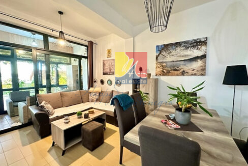 dining room view with dining table and a plant on it, sofa with coffee table behind the table and big window with green plants and sun